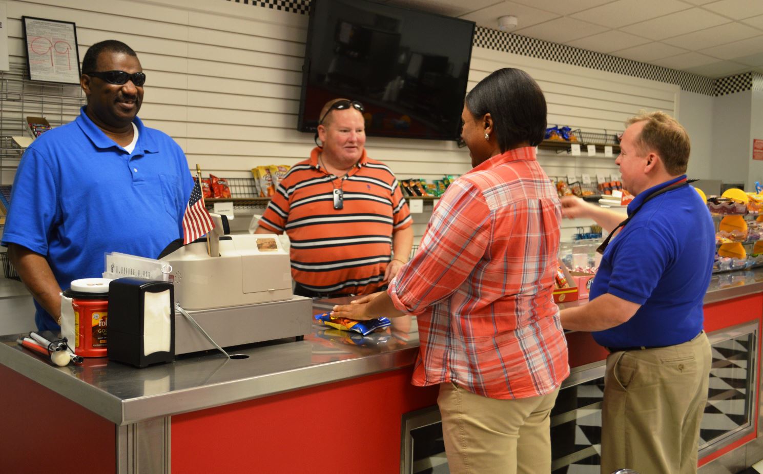 Four subjects in food vending counter smiling and talking with each other: the adult African American male working the cash register is blind. He is behind the counter with an adult white male coworker and both are speaking with two customers, an adult African American female and an adult white male.