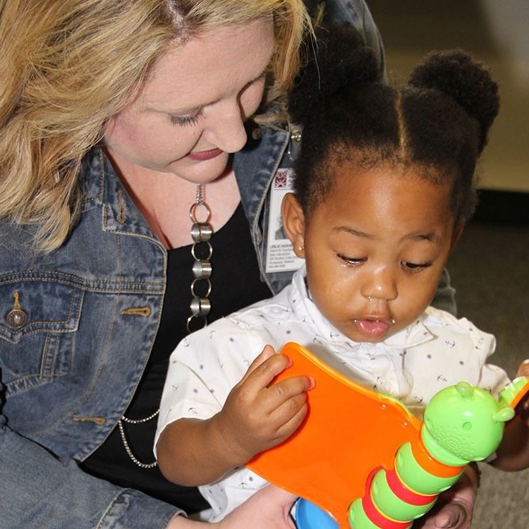 Adult female with a female toddler sitting in her lap reading a toy book together