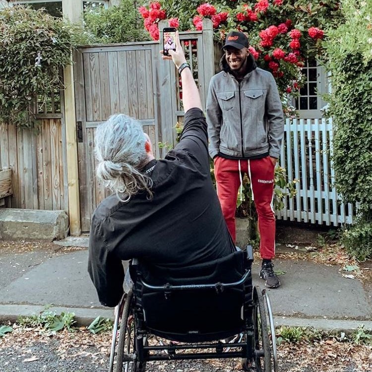 Back of adult, elderly male sitting in a wheelchair reaching up with right hand with forward-facing adult male smiling and a flower bush behind him